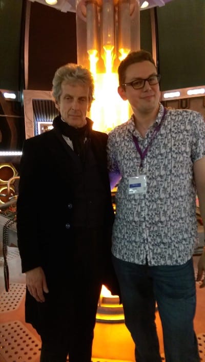 Peter Capaldi and Cavan Scott on the TARDIS set in 2016, in the control room with the glowing time rotor behind them. Capaldi wears his Doctor costume.